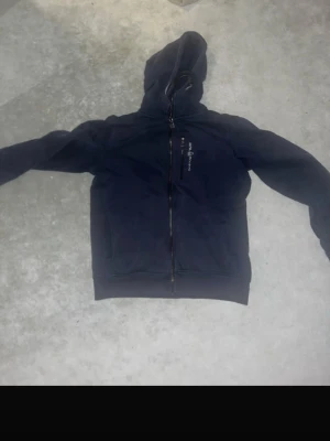 Mörkblå zip-hoodie från sail racing  - Säljer en mörkblå hoodie med dragkedja från Peak Performance. Tröjan har huva med logga baktill och två fickor framtill. Perfekt för en avslappnad och sportig stil. Materialet är mjukt och bekvämt, och passformen är klassisk med ribbade muddar.