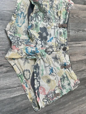 Färgglada blommiga shorts med knappar - Snygga shorts med blommigt mönster i flera färger som blått, grönt, rosa och svart på ljus botten. Shortsen har knappar framtill och dekorativa detaljer. Perfekta för varma dagar och en avslappnad stil.