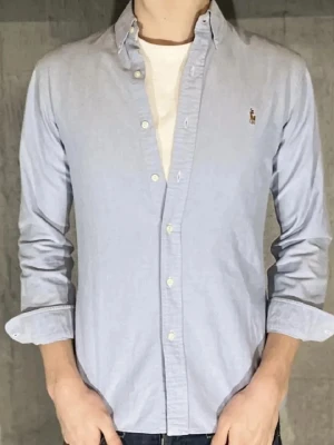 Ljusblå skjorta från Polo Ralph Lauren - Klassisk ljusblå skjorta från Polo Ralph Lauren med button-down krage och broderad logga på bröstet. Skjortan har vita knappar och är perfekt för en stilren look.