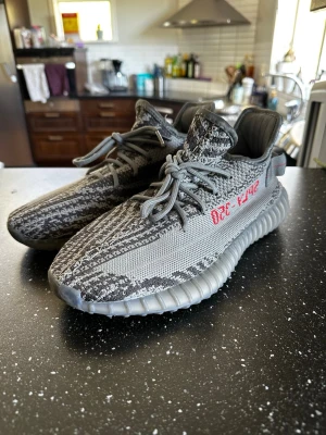 Adidas Yeezy Boost 350 V2 Beluga - Grymma Adidas Yeezy Boost 350 V2 Beluga med grå och mörkgrå mönstrad ovandel i mesh och textil. Sidan har den ikoniska röda SPLY-350-texten. Skorna har en tjock, räfflad sula i gummi och bekväm Boost-dämpning. Perfekta för dig som gillar streetwear och sneakerskultur.