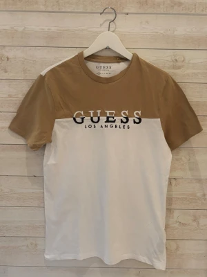 Beige och vit t-shirt från Guess - Snygg t-shirt från Guess med beige ovandel och vit nedre del. Stor logga 'GUESS LOS ANGELES' broderad över bröstet. Klassisk rund hals och korta ärmar. Tillverkad i mjuk bomull för en skön känsla.