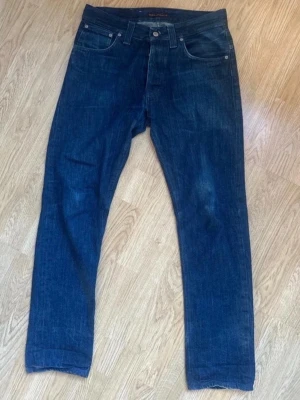 Mörkblå jeans från Nudie Jeans - Säljer ett par klassiska mörkblå jeans från Nudie Jeans med raka ben och femficksdesign. Jeansen har en snygg denimfinish och detaljer som metallknappar och läderpatch bak. Perfekt för dig som gillar stilrena och tidlösa jeans.