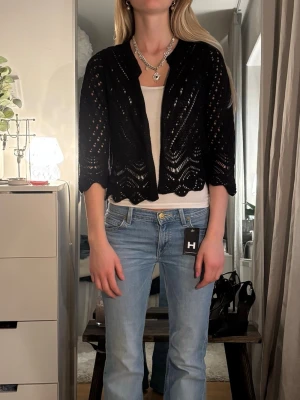 Svart virkad kofta från Hunkydory - Snygg svart virkad kofta från Hunkydory med kortare passform och vågig kant nertill. Koftan har ett öppet mönster och trekvartsärmar, perfekt att slänga över ett linne eller t-shirt för en chill look.