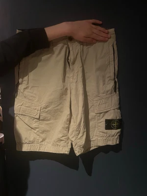 Beige cargoshorts från Stone Island - Säljer ett par beige cargoshorts från Stone Island med klassisk logotyp-patch på benet. Shortsen har flera fickor och stängs med knapp och dragkedja. Perfekta för sommaren och har en avslappnad passform.