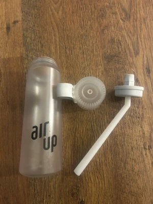 Air Up vattenflaska med sugrör - Transparent vattenflaska från Air Up med svart logga, vit skruvkork och vitt sugrör. Flaskan är tillverkad i plast och har en modern, avlång form. Perfekt för att smaksätta vatten med doftpoddar. Knappt använd men noggrant tvättad ändå