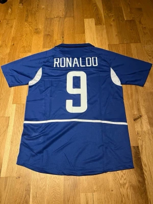 Brazil 2002 Ronaldo #9 - Snygg blå fotbollströja från Nike med Brasilien-emblem och nummer 9 på både fram- och baksida. RONALDO står tryckt på ryggen. Tröjan har vita detaljer på sidorna och runt numret. Perfekt för dig som älskar fotboll och vill hylla en legend.