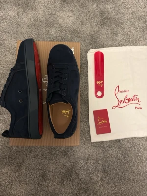 Mörkblå sneakers från Louboutin - Snygga mörkblå sneakers från Christian Louboutin i mocka med klassisk röd sula. Lågt skaft, svarta skosnören och diskret logga på insidan. Perfekta för dig som vill ha en stilren och exklusiv look med en ikonisk touch.