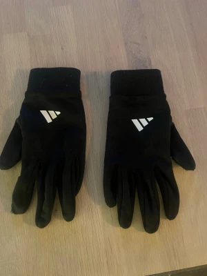 Svarta sporthandskar med greppmönster. Perfekt för fotbollsspelare. - Svarta sporthandskar med vitt greppmönster på handflatan och vitt tryck på ovansidan. Handskarna har ribbade muddar och är tillverkade i ett syntetmaterial som passar perfekt för utomhusaktiviteter under kyligare dagar. Perfekt för fotboll.