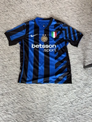 Inter Milan x Nike matchtröja  - ALDRIG ANVÄND. Snygg Inter Milan matchtröja i blått och svart med grafiskt mönster, klubbmärke och sponsortryck på bröstet. Kortärmad modell i lätt och ventilerande material, perfekt för fotboll eller träning. Klassisk V-ringning och gula detaljer på sidorna.