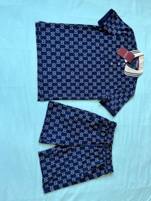 Mörkblått set från Gucci med GG-mönster - Snyggt tvådelat set från Gucci i mörkblått med klassiskt vitt GG-mönster över hela plagget. Setet består av en kortärmad pikétröja med vit krage och matchande shorts med resår i midjan. Perfekt för en trendig och avslappnad look.