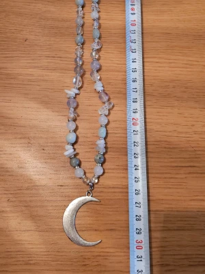 Halsband med pärlor och månhänge - Unikt halsband med halvädelstenar i pastellfärger och transparenta toner. Halsbandet har ett stort månhänge i silverfärgad metall som ger en cool och mystisk vibe. Perfekt accessoar för dig som gillar statement-smycken och boho-stil.