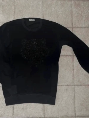 Svart Kenzo sweatshirt med tiger - Säljer en svart sweatshirt från Kenzo med det ikoniska tigerhuvudet broderat på bröstet. Tröjan har rund halsringning, ribbade muddar och är tillverkad i mjukt bomullsmaterial. Perfekt för dig som gillar streetwear och vill ha något stilrent.passar som S och M !