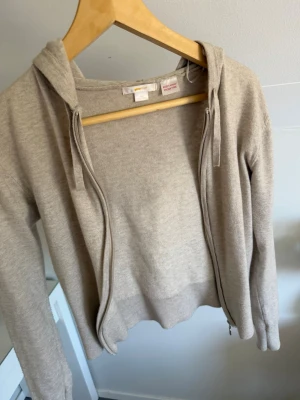 Beige hoodie/kofta från Gina Tricot - Snygg beige hoodie från Gina med dragkedja framtill och huva med snören. Tillverkad i en mjuk och skön bomullsmix som passar perfekt till chill dagar. Långa ärmar och ribbade muddar ger en clean look. Har knappt använts då den kom med ett litet hål på armen