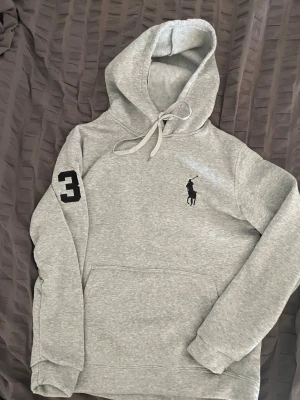 Grå hoodie från Polo Ralph Lauren - Snygg grå hoodie från Polo Ralph Lauren med svart broderad logga på bröstet och en stor svart siffra 3 på ärmen. Klassisk känguruficka och dragsko i huvan. Perfekt för en chill och sportig stil.