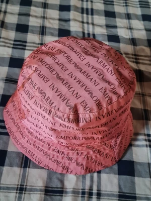 Rosa bucket hat från Emporio Armani - Cool rosa bucket hat från Emporio Armani med logotyptryck över hela hatten. Tillverkad i ett lätt tyg som känns skönt på huvudet. Perfekt accessoar för att lyfta din outfit med en trendig touch.