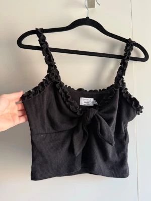 Svart croppad linnetopp med volang - Supersöt svart croppad linnetopp från Pamela x NA-KD med volangdetaljer längs axelband och urringning. Toppen har knyt framtill och är tillverkad i ribbad bomull. Perfekt för dig som gillar trendiga och feminina plagg. Katt finns i hemmet!