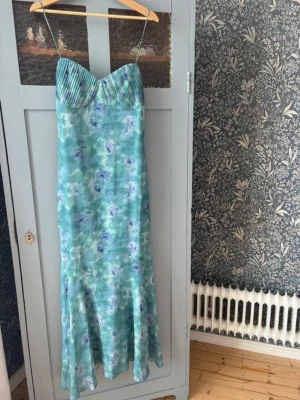 Blommig långklänning med tunna band - Ljusblå långklänning med blommigt mönster och tunna axelband. Klänningen har ett plisserat liv och en mjukt utsvängd nederdel. Materialet ser ut att vara glansigt, troligen satin eller polyester, vilket ger en elegant känsla. Perfekt för sommarens alla tillfällen.