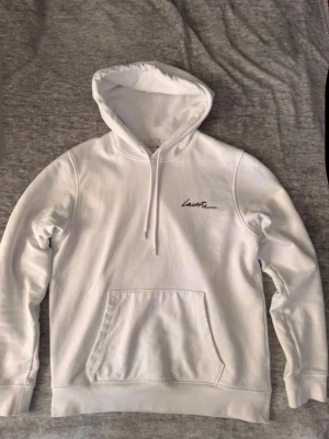 Lacoste hoodie - Snygg vit hoodie från Lacoste med broderad krokodil på ärmen och diskret Lacoste-signatur på bröstet. Klassisk känguruficka framtill och justerbar huva med dragsko. Tillverkad i mjuk bomullsblandning för skön känsla och stilren look riktigt fet nu till sommaren.