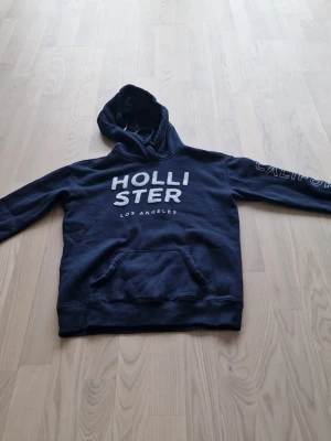 Mörkblå hoodie från Hollister - Mörkblå hoodie från Hollister med vit text på bröstet och tryck på ena ärmen. Klassisk känguruficka framtill och huva. Perfekt för en avslappnad och cool stil. Mjuk bomullsblandning som känns skön mot huden.