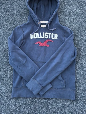 Blå hoodie från Hollister - Mörkblå hoodie från Hollister med broderad vit logga och röd fågel på bröstet. Klassisk känguruficka framtill och justerbar huva med dragsko. Mjuk bomullskänsla och avslappnad passform, perfekt för en chill stil.