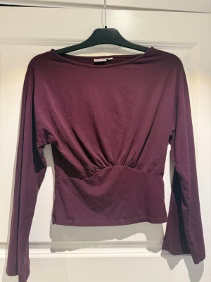 Vinröd långärmad topp med rynk - Vinröd långärmad topp med rund halsringning och snygg rynkdetalj framtill i midjan. Modellen är figurnära och har en enkel, stilren design som passar till många olika looks.
