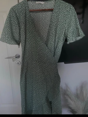 Grön omlottklänning från ONLY - Snygg ljusgrön omlottklänning från ONLY med svart prickigt mönster. Klänningen har korta ärmar, volangkant nertill och knytband i midjan. Perfekt för sommarens alla tillfällen!