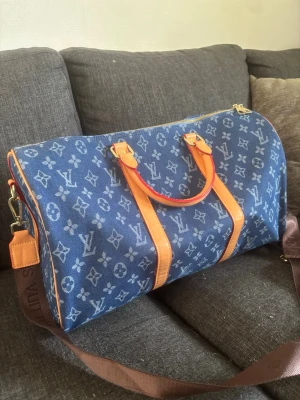 Louis Vuitton duffle bag - Säljer en snygg blå weekendbag från Louis Vuitton i denimtyg med klassiskt monogrammönster. Väskan har bruna läderdetaljer, guldiga metalldelar och en bred axelrem i brunt tyg. Perfekt för resor när du vill sticka ut med stil.