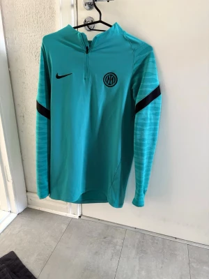 Turkos Nike Inter Milan träningströja - Snygg turkos långärmad träningströja från Nike med Inter Milan-logga på bröstet. Tröjan har halv dragkedja, svarta detaljer på ärmarna och är tillverkad i Dri-Fit-material som andas. Perfekt för fotbollsträning eller gymmet.