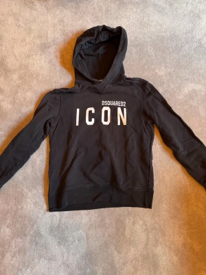 Svart hoodie DSQUARED2. - Svart hoodie från DSQUARED2 med stor vit text 'ICON' på bröstet och mindre logga ovanför. Klassisk modell med huva och ribbade muddar. Ny pris 2500kr.