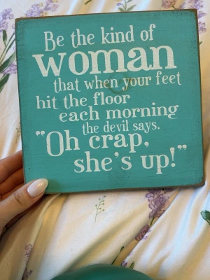 Turkos träskylt med inspirerande citat - Dekorativ fyrkantig träskylt i turkos med vitt tryck: 'Be the kind of woman that when your feet hit the floor each morning the devil says: Oh crap, she's up!'. Skylten har ett handtag på baksidan för enkel upphängning.
