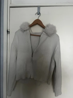 Vit stickad hoodie med fuskpäls - Mysig vit stickad hoodie med dragkedja och huva kantad med fluffig fuskpäls. Tröjan har ribbstickade muddar och en avslappnad passform, perfekt för kalla dagar. Enkel och stilren design som passar till många outfits.