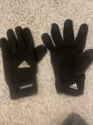 Svarta fleecehandskar från Adidas - Svarta fleecehandskar från Adidas med AEROREADY-tryck och kardborreknäppning vid handleden. Perfekta för kyliga dagar och har halkskydd på fingertopparna. Klassisk Adidas-logga på ovansidan.