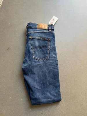 Blåa jeans från Nudie  - Snygga blå jeans från Nudie Jeans med klassisk femficksdesign och läderpatch bak i midjan. Jenasen jar en riktigt snygg tvätt och en liten slitning på knät. Hålet är nyss lagat eftersom det är en lapp på Jeansen. Hör gärna av er vid minsta fundering om plagget.!😊