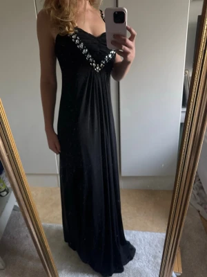 Svart långklänning med strassdetaljer - Elegant svart långklänning med smala axelband och v-ringning dekorerad med glittrande strass. Klänningen har en figurnära passform upptill och faller snyggt längs kroppen. Perfekt för speciella tillfällen så som bal eller fest.