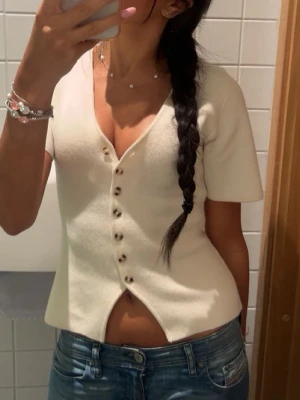 Vit kortärmad topp med knappar - Snygg vit topp med korta ärmar och v-ringning. Toppen har dekorativa knappar framtill och en lätt utsvängd passform med en liten slits nedtill. Perfekt till jeans eller kjol för en trendig look.