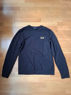 Svart sweatshirt EA7 Emporio Armani - Stilren svart sweatshirt från EA7 Emporio Armani med guldfärgad logga. Tröjan är i storlek L men passar mer till M eller S. I princip fortfarande ny, testat den endast 2 gånger.