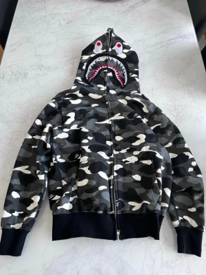 BAPE camo shark hoodie - Säljer en ikonisk BAPE hoodie med grå, svart och vit camomönster. Huvan har det klassiska hajansiktet med tänder, ögon och dragkedja hela vägen upp. Ribbstickade muddar och midja, samt två fickor fram. Perfekt för dig som gillar streetwear och vill sticka ut.
