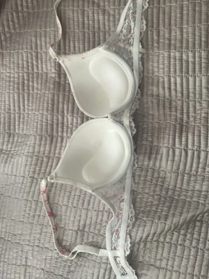 Vit bh med rosa rosetter och spets - Söt vit bh med rosa rosett-mönster och dekorativ vit spets längs kanterna. Bh:n har vadderade kupor och justerbara axelband. Materialet är mjukt och bekvämt, perfekt för dig som gillar en feminin och lekfull stil. Priset går att diskutera. 