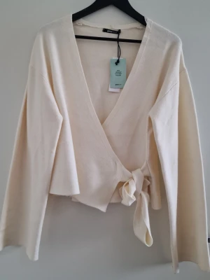 Krämvit omlott tröja från Gina Tricot - Snygg krämvit omlott stickad tröja från Gina Tricot med breda ärmar och knytband i midjan. Koftan har djup v-ringning och är gjord i mjukt stickat material. Perfekt att styla över ett linne eller topp för en trendig look. Storlek L.