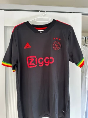 Ajax svart matchtröja Adidas - Snygg svart Ajax matchtröja från Adidas med Ziggo-logga på bröstet. Tröjan har röda, gula och gröna detaljer på axlar och ärmslut samt Ajax-emblem och Adidas-logga i rött. Tillverkad i lätt och ventilerande material, perfekt för fotboll eller träning.