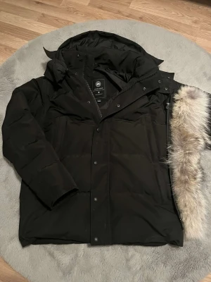 Svart dunjacka från Canada Goose - Snygg svart dunjacka från Canada Goose med avtagbar huva och en fluffig pälskant. Jackan har dragkedja och knappar framtill samt två stora fickor. Perfekt för kalla vinterdagar och riktigt varm tack vare dunfyllningen.