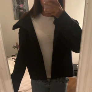 Marinblå dubbelknäppt rock med bred krage - Snygg svart rock med bred krage och dubbelknäppning framtill. Kortare modell som ger en modern look. Passar perfekt till jeans och har en klassisk, stilren design. Materialet ser ut att vara ull eller ullblandning.