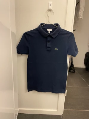 Marinblå pikétröja från Lacoste - Klassisk marinblå pikétröja från Lacoste med korta ärmar och den ikoniska krokodilloggan på bröstet. Tröjan har krage och knappar framtill, perfekt för en clean och stilren look. Tillverkad i mjuk bomull som känns skön mot huden. Junior storlek 14 år. 