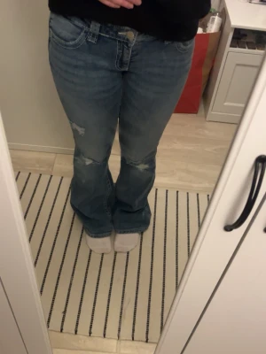 Blå bootcut jeans från Gina 14+ - Säljer ett par blå bootcut jeans med slitningar och snygga detaljer på bakfickan. Jeansen har låg midja och klassisk femficksmodell. 