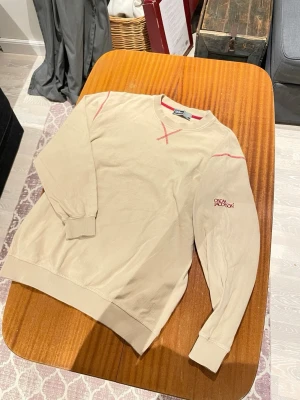 Oscar Jacobson Beige Sweatshirt - Stilren beige sweatshirt från Oscar Jacobson med röda kontrastsömmar vid halsringningen och ärmslut. Den är använd få gånger. Storleken är L men den passar som M (väldigt liten i storleken