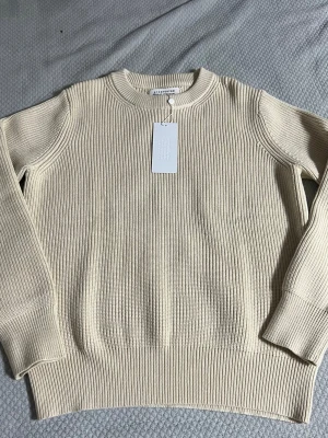 Beige stickad tröja från Maison Margiela - Snygg beige stickad tröja från Maison Margiela med ribbad struktur och rund halsringning. Tröjan har långa ärmar och breda muddar vid ärmslut och nederkant. Perfekt för dig som gillar minimalistisk och stilren design.