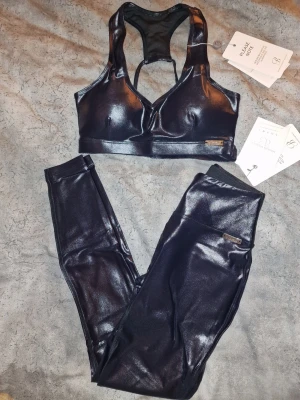 Bombshell Sportswear Träningsset - Sport-bh och Leggings - Träningsset med sport-bh och matchande leggings. Setet har en cool wet look-effekt, sport-bhn har breda axelband och meshdetaljer i ryggen. Skriv för fler bilder.  *Supersnyggt till gymmet, dansen eller med en sweatshirt för powerwalken  *Du får hela setet till priset utav leggingsen