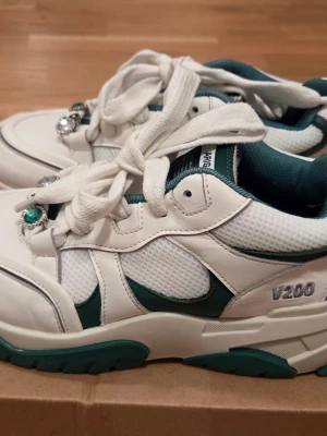 Axel Arigato V200 vita sneakers - Säljer ett par vita Axel Arigato V200 sneakers med gröna detaljer och meshpaneler. Skorna har chunky sula, breda vita skosnören och dekorativa smycken med yin yang och gröna stenar. Märket syns på plösen och sidan. Perfekta för dig som gillar statement sneakers.