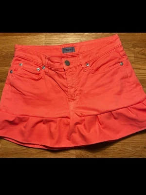 Korallrosa kjol med volang från Filippa K - Snygg kort kjol i korallrosa från Filippa K. Kjolen har volang nertill, klassiska jeansfickor och knapp framtill. Perfekt för dig som vill ha en färgstark och trendig look.