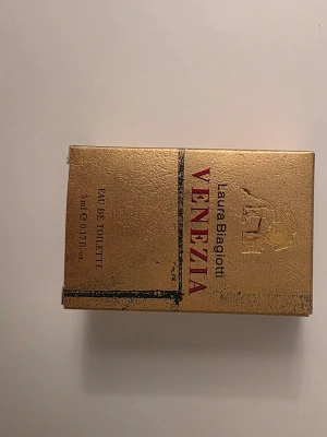 Laura Biagiotti Venezia Eau de Toilette - Laura Biagiotti Venezia Eau de Toilette i en elegant miniflaska med guldfärgad kartong. Flaskan har en unik form med vinröd topp och genomskinligt glas som visar den gyllene vätskan. Perfekt för dig som gillar exklusiva och klassiska dofter.